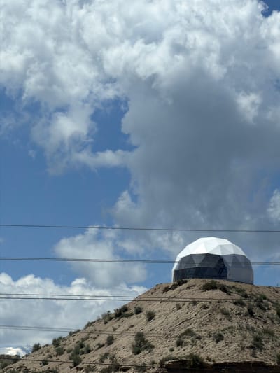 A geodesic dome structure sits atop a dry, rocky hill under a partly cloudy sky in Tong, Kyrgyzstan. The dome, composed of white triangular panels, has a transparent lower section that reveals a dark interior. Several power lines are strung across the frame, bisecting the view of the dome and the hill. The hill is sparsely covered with dry vegetation and shows signs of erosion. In the distance, a snow-capped mountain range is visible on the left. The overall scene suggests a remote, arid environment.