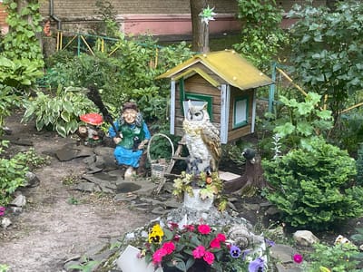 The image depicts a garden scene with several decorative elements. In the foreground, a pathway made of dirt and stones winds through the garden. To the left, a gnome-like figure wearing a blue dress and green vest, holding a broom and a basket, is situated among plants and rocks. Beside the gnome, there are small mushroom figures. A detailed owl sculpture stands near a small wooden house with a yellow roof. In front of the owl sculpture, a cluster of pink and yellow flowers blooms, accompanied by a decorative snail sculpture and other floral elements. To the right of the owl, a small black bird sculpture perches on a tree stump, adjacent to a lush green bush. The background features a brick building and a green fence with yellow and blue accents. The overall setting appears to be a residential garden in the city of Chornoye, Russia.