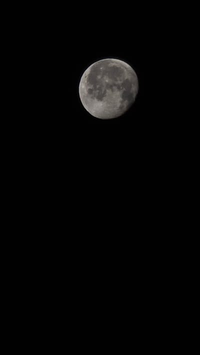 The image shows a close-up view of the moon against a black, starless sky. The moon is partially illuminated, showing craters and surface features. The bottom portion of the image is entirely black. There is no indication of the city of Tatabánya, Hungary, or any urban elements in the picture.