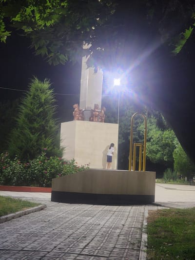 A young girl with dark hair stands with her back to the camera, wearing a white t-shirt and a blue skirt. She is positioned on a raised platform in front of a tall, light-colored monument. The monument has bronze sculptures of mythological creatures at its top. To the left of the monument is a tall, cone-shaped evergreen tree, and in front of that is a bed of flowering plants with pink and orange blossoms. To the right of the girl and the monument is a yellow, abstract sculpture. The scene is illuminated by a bright light source positioned above and to the right of the monument, causing lens flare. The ground in front of the monument is paved with interlocking gray tiles. In the background, there are more trees and foliage. The overall setting appears to be a park or public square in Jetisay, Kazakhstan, likely in the evening or at night due to the dark sky and artificial lighting.