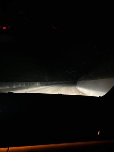 The image depicts a nighttime view from inside a vehicle traveling on a dark, unlit road, likely in the vicinity of Byala Reka, Bulgaria. The headlights of the car illuminate a section of the road ahead, revealing a paved surface with a slight curve to the right. On either side of the road, dense, dark foliage is visible, with some hints of a low concrete barrier along the left side. The sky is completely dark, indicating it is nighttime. The interior of the car is largely obscured by shadow, though a segment of the dashboard is visible in the foreground, with a faint orange light emanating from a control panel on the right side. The overall impression is one of solitary travel through a rural or forested area at night.