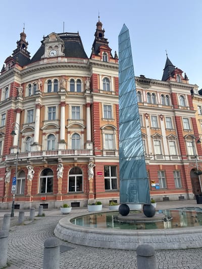 The image depicts a fountain with a modern glass obelisk sculpture in the foreground, set against the backdrop of an ornate, historic building. The fountain's basin is circular and constructed of stone, with water reflecting the sky and the surrounding architecture. The glass obelisk is tall and slender, composed of multiple panels with a rippled, reflective surface, and rests on three black spherical bases. The building behind the fountain is multi-story with a red and cream facade, featuring arched windows, decorative columns, balconies, and elaborate architectural detailing. A clock is visible on the building's roofline, which appears to be Roman numerals, indicating MDCCCXIV. The ground surrounding the fountain is paved with cobblestones, and there are several concrete bollards scattered around. In the background, the sky is clear and pale blue, suggesting it is either early morning or late afternoon. A street lamp with a hanging basket of flowers is also visible to the left of the building. The scene appears to be a public square or plaza in a European city. Based on the architecture and the visible signage, this location is identified as the Fountain with the glass obelisk, located in Karlovy Vary, Czechia.