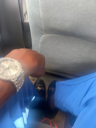A partial view of an individual is depicted from a first-person perspective inside a vehicle in Elimgbu, Nigeria. The subject's dark-skinned left wrist, visible on the left, is adorned with a large, circular, silver-toned wristwatch featuring extensive surface embellishments. The individual wears light blue trousers and black loafer-style shoes, each detailed with a prominent gold-colored metal bar across the vamp. A red-orange strap from an unseen object is partially visible on the vehicle's floor between the subject's feet. The vehicle interior includes a light grey fabric car seat, visible directly in front and to the right, and a dark grey interior panel on the far left. The scene is evenly lit.