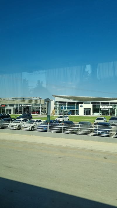 A vertical, eye-level shot shows a modern car dealership with a clear blue sky in the background. The dealership features large glass windows and a curved, contemporary roofline. Several cars are parked in front of the building, both on the lot and in a display area separated by a low metal fence. The dealership's signage includes "Kapsiotis" and a Volkswagen logo. Reflections of trees and buildings are visible in the glass of the dealership and the car windows. The foreground shows a paved road and sidewalk. The image is a photograph taken from a slight distance, possibly from another vehicle or a slightly elevated position. The overall scene suggests a commercial area on a bright, sunny day.