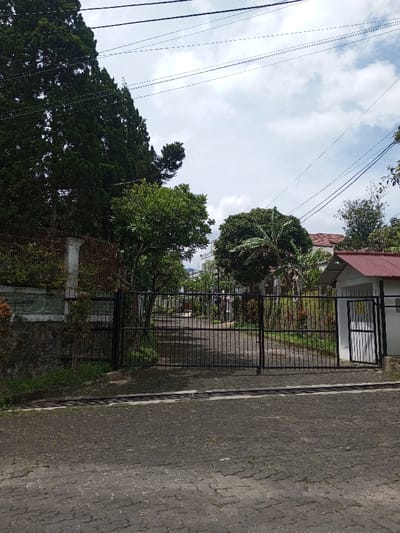 The scene depicts a gated entrance in Vieng Phouka, Laos, under an overcast sky. In the immediate foreground, a dark gray paved surface, constructed from interlocking pavers, extends across the frame, showing minor discoloration and scattered debris. A long, dark metal grate runs horizontally across this pavement, just before a set of gates.
A large, black metal double gate, composed of vertical bars and horizontal cross-members, stands closed in the midground, serving as the primary vehicular entrance. To the left of this main gate, a multi-layered barrier consists of a rough-hewn stone base, topped by a painted concrete wall, and further capped with a section of rusty, dark metal fencing. Tall, dark green coniferous trees rise significantly above this wall on the far left, extending into the upper left quadrant of the image.
To the right of the main gate, a small, white, single-story building with a distinctive red corrugated metal roof is visible. A smaller, black metal pedestrian gate, matching the design of the main gate, is partially open, revealing a glimpse inside this structure.
Beyond the main gate, a paved roadway or driveway extends into the background, flanked on both sides by dense, green vegetation, including broadleaf trees, shrubs, and at least one banana plant visible on the right side of the drive. Multiple residential structures are discernible further down the drive, including one with a reddish-orange tiled roof on the right and a white-sided building further back on the left.
The upper portion of the image is dominated by an overcast sky, with shades of white and gray clouds. Multiple black utility lines traverse the sky horizontally across the upper frame. No individuals or active interactions are present within the frame.