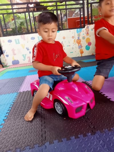 The image captures a play area in Kuwukan, Indonesia. A young male child, approximately 2-4 years old, is seated on a bright pink, low-profile ride-on toy car, holding its black steering wheel with both hands. He wears a red short-sleeved t-shirt with "LEVI'S" branding on the sleeve stripe, blue denim shorts, and is barefoot. His gaze is directed slightly downwards and forward. To the right, a portion of another child is visible, wearing a red t-shirt and dark shorts, kneeling on a mat. The floor is covered with interlocking foam mats in various colors, including blue, green, purple, and dark grey, featuring a textured surface. The background includes a light-colored wall decorated with colorful child-themed illustrations, such as letters and cartoon figures, surmounted by a dark metal railing, beyond which some green foliage is discernible. The setting suggests an indoor or semi-outdoor children's play facility.