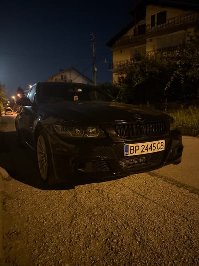 A black BMW sedan is parked on a gravel shoulder beside a paved road at night. The car's headlights are on, illuminating the license plate which reads "BP 2445 CB". The background shows a residential building with balconies and trees. The scene is likely in Belene, Bulgaria, given the license plate format.