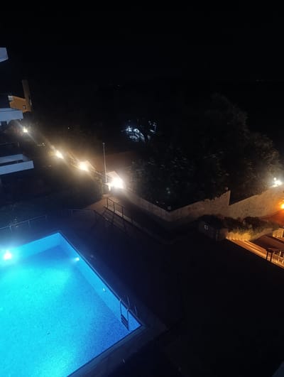 An outdoor swimming pool illuminated with blue lights dominates the foreground of the image. To the right of the pool, a dark patio area with railings is visible. Beyond the patio, a walkway with stairs and railings is illuminated by warm, yellow lights, leading past a stone wall and lush green vegetation. In the background, several buildings with red-tiled roofs are visible, also lit by artificial lights. The scene is set at night, with the dark sky above. The distant horizon suggests the presence of water, possibly the sea, with faint lights indicating a distant shoreline. The location is identified as Urla, Turkey.