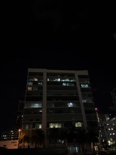 A tall, modern building dominates the right side of the frame against a stark, black night sky. The building features a grid-like facade of glass windows, some illuminated with interior lights. Its architectural design is linear and angular. Palm trees partially obscure sections of the building. Another building with many windows is visible on the right, further back. A purple neon X is seen atop one of the buildings.