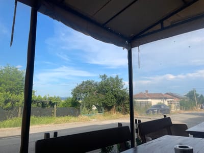 The image presents a daytime view from inside a covered outdoor seating area in Konstantinovo, Bulgaria, looking out onto a rural or semi-urban street. The immediate foreground reveals dark wooden tables and benches, with a metallic ashtray positioned on the table to the lower right. Overhead, a beige fabric canopy is supported by dark metal structural elements, including posts framing the view through a clear plastic or glass barrier. This barrier exhibits slight reflections and smudges.
Beyond the barrier, a narrow road or path extends horizontally across the midground. To the left, a green wire fence is partially concealed by overgrown green grass and weeds. Behind the fence, a structure draped in vines and additional trees are visible. Further to the right, a large green metal gate is partially obscured by dense foliage. A dark-colored Audi vehicle is parked on the right side of the road. Behind the vehicle, an aged, single-story house with a distinct red-tiled roof and weathered facade is present. A portion of another similar house and a utility pole are visible further to the right. The background features a distant tree-lined horizon beneath a clear blue sky with scattered white clouds. No people or specific actions are depicted in the scene.