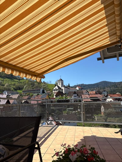 A view from a balcony overlooks Forbach, Germany. The town features residential buildings with red-tiled roofs and a church with a clock tower. Behind the town is a forested hillside. A tan and white striped awning extends from the left of the frame. The balcony itself is tiled with light brown square tiles. A metal railing encloses the balcony, and a portion of a dark chair is visible in the foreground. Flowers are planted near the edge of the balcony on the right. The sky is clear and blue.