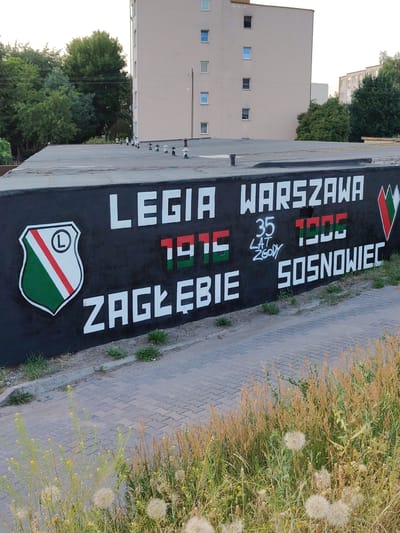 This media file captures a mural painted on a black, rough-textured wall. The mural features prominent text and graphics related to football clubs.
The central subject of the mural is the text "LEGIA WARSZAWA" in large, white capital letters. To the left of this text is a shield emblem with a "L" inside a circle, and the shield is divided into sections of white, red, and green. Below "LEGIA WARSZAWA," there are two sets of numbers in red and green, representing years. The first set, "1915," is in red and green. To its right, the text "35 LAT ZGODY" is written in smaller, white script, with "35" and "ZGODY" in white and "LAT" in grey. The second set of numbers, "1905," is in green. Below these numbers, in large white capital letters, is the word "ZAGŁĘBIE" and to the right of that, "SOSNOWIEC".
The scene is outdoors, with a paved walkway or road in the foreground. Behind the wall, a light-colored, multi-story building is visible. The setting appears to be urban or semi-urban, with trees and foliage in the background. The time of day seems to be daytime, possibly late afternoon or early evening, given the lighting conditions. The weather appears to be fair.
The mural likely represents a sports rivalry or alliance between the football clubs Legia Warsaw and Zagłębie Sosnowiec, with "35 LAT ZGODY" translating to "35 Years of Harmony" or "35 Years of Friendship," suggesting a significant historical connection between the clubs. The date 1905 might refer to the founding year of one of the clubs, and 1915 to the other. The location context provided is Nowy Dwór Mazowiecki, Poland.