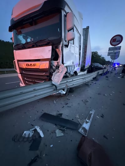 The image depicts the aftermath of a multi-vehicle collision on a highway in Chirpan, Bulgaria, observed at dusk or nighttime. A white DAF semi-truck is positioned prominently in the foreground, facing the camera at an angle. Its front end exhibits severe damage, with the bumper, grille, and headlights crushed and fragmented. The DAF logo on the grille is still visible amidst the deformation. The truck has impacted and extensively damaged a metal guardrail, which is bent inwards and broken at the point of impact.
Scattered on the asphalt road surface around the truck and guardrail are numerous pieces of vehicle debris, including plastic fragments, metal components, and a coiled spring. Further down the road, behind the damaged semi-truck, a dark-colored passenger vehicle is visible, also appearing to be damaged. Blue flashing lights, consistent with emergency services (police or ambulance), are discernible in the distance, illuminating the scene. Several individuals are present in the background near the emergency lights and the damaged passenger vehicle, though their specific actions are not discernible.
Two road signs are visible on the right side of the road. The upper sign is a circular red and white regulatory sign depicting a truck and a car, indicating a restriction, possibly "no overtaking for trucks." Below it, a rectangular white sign displays "↑ 2600M ↑" and "7:00 – 19:00", suggesting a time-based restriction (between 7 AM and 7 PM) applicable over a distance of 2600 meters. The sky in the background is a deep twilight blue, and a line of trees is visible along the roadside.