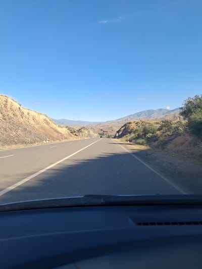 The image shows a road winding through a mountainous landscape, likely taken from the perspective of a vehicle. The road is paved with a white line marking the lanes. To the left of the road, a steep, rocky mountainside covered in sparse vegetation rises towards a clear blue sky. In the distance, more mountains are visible, shrouded in a light haze. To the right of the road, a dark blue object, possibly the interior of a vehicle, is in the foreground. The sunlight suggests it is daytime, and the clear sky indicates fair weather. The landscape appears arid and rugged. The provided location context is Vayk, Armenia. There is no visible text in the image.