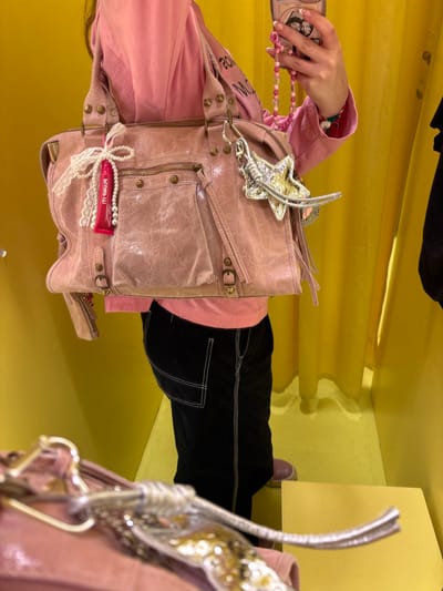 This image depicts a partial view of a person within an indoor setting, likely a changing room or retail fitting area, located in Leganés, Spain. The environment features uniform bright yellow walls and floor, with vertical drapery visible in the background. A small yellow cuboid object rests on the floor in the lower right. The subject, a person whose face is not visible, is wearing a pink long-sleeved shirt, black trousers with white contrast stitching, and light pink shoes.

The person is holding a large, light pink handbag, appearing to be made of distressed leather, equipped with multiple external pockets, zippers, and metallic stud details. Attached to the left handle of the handbag is a decorative assembly consisting of a white lace bow, a string of white pearls, and a small red cylindrical object with the text "PLUMP IT!" in white. A prominent silver-colored keychain, shaped like an ornate star with additional metallic feather-like elements, is attached to the right side of the handbag near its handle.

The subject's right hand is visible, holding a smartphone with a light-colored case. A pink beaded strap hangs from the smartphone. A green and red beaded bracelet is visible on the subject's right wrist. A blurry reflection of the handbag and keychain is visible in the lower foreground, suggesting the presence of a mirror directly in front of the subject.