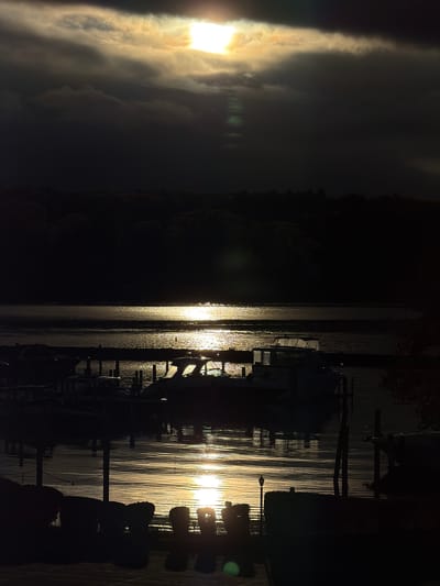 The image depicts a lakeside scene at dusk, possibly in Laconia, USA. A bright sun is obscured by clouds on the left, casting a strong glare. The water surface reflects the sunlight. A dark shoreline is visible in the middle. On the right, silhouetted docks and boats are present, with their forms reflected in the water. The overall scene suggests a calm evening at a marina or lakeside location.