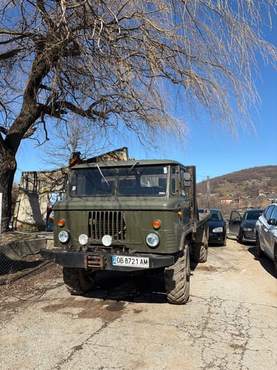An olive green flatbed truck, identified by its Bulgarian license plate "OB 8721 AM," is parked on a cracked asphalt surface in Babintsi, Bulgaria. The truck features a prominent front grille, two main headlights, two amber turn signals, and three circular auxiliary lights mounted on a heavy-duty black bumper with a central winch mechanism. Its large, knobby off-road tires show signs of dirt and mud. The cab has two windshield wipers and orange marker lights on the roof.

To the left of the truck, a large, bare deciduous tree with thick branches dominates the upper left frame. Behind the tree, a light-colored building with a textured wall is visible, casting shadows. To the right of the truck, a dirt or gravel road recedes into the background, where a black sedan with its front passenger door open, another dark sedan, and a silver SUV are parked.

In the distant background, rolling hills or mountains are visible under a clear, bright blue sky. Several houses with red or orange roofs are scattered on the lower slopes of these hills. No people are present in the scene. The strong sunlight casts distinct shadows across the ground and buildings.