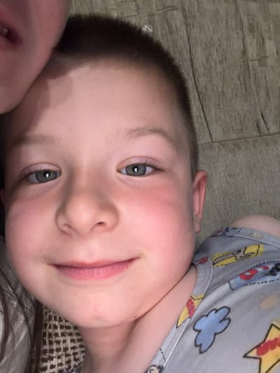 The image is a close-up, low-angle selfie of a young boy. He has fair skin, light brown hair cut short, and grey-blue eyes. His left cheek is flushed, and he is wearing a light grey shirt with a colorful cartoon pattern of rockets and clouds. Part of another person's face is visible in the upper left corner of the frame. The background appears to be a textured surface, possibly upholstery, with a pattern of vertical lines. The scene is indoors, likely a domestic setting. The image is not located in the city of Zmeitsa, Bulgaria.