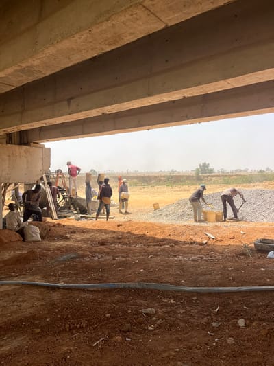 The image captures a construction site beneath a concrete overpass or bridge structure in Wuciciri, Nigeria. Approximately ten male laborers are visible, engaged in various tasks on disturbed reddish-brown earth under a hazy sky. On the left, a group operates an orange-framed cement mixer; one individual stands elevated on a platform next to the mixer, while others stand nearby, some carrying white sacks on their heads or interacting with the equipment. In the right mid-ground, two men are actively shoveling grey aggregate from a large pile into yellow buckets. Other workers are distributed across the scene, some carrying materials. A blue hose or pipe extends across the immediate foreground. The background consists of flat terrain with sparse vegetation.