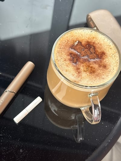 A top-down, medium shot shows a glass mug of frothy coffee with a sprinkle of cocoa powder on top, positioned on a dark, reflective surface, likely a table. To the left of the mug, there is a golden electronic smoking device and a white cigarette stick with an orange tip. In the background, a portion of a similar electronic device is visible. The scene appears to be indoors, likely in a domestic or public setting within the city of Semiluki, Russia. The lighting suggests it is daytime.