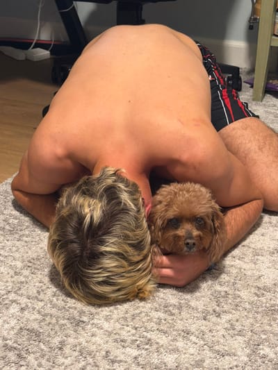 An aerial view shows a shirtless, fair-skinned man with blond hair on a gray carpet. He is lying on his side with his arms wrapped around a small, brown dog. The dog looks up at the camera. The man's back is visible, and he is wearing red, white, and black shorts. A desk or shelving unit is partially visible in the background.