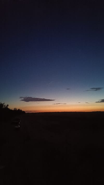 A vertical, low-angle shot captures a twilight landscape in Martynivs'ke, Ukraine. The sky transitions from deep blue at the top to a gradient of lighter blue, then orange, and finally a thin band of yellow just above the horizon. wispy clouds are scattered across the sky. The foreground is a dark, silhouetted expanse of land, suggesting a rural or undeveloped area. On the left side of the frame, the dark silhouette of trees is visible, and to the left of center, the dark shape of a vehicle, possibly a car, is partially obscured. The overall impression is one of dusk or the very early stages of nightfall in a quiet, open environment.