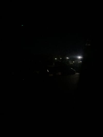 The image depicts a nighttime scene in a dark outdoor environment, possibly a parking lot or street. A faint green light is visible in the upper left quadrant. In the mid-ground, the silhouette of a car is discernible, with its headlights faintly illuminated. Several streetlights are visible in the background, casting a soft glow on the surroundings. The overall impression is one of low visibility, typical of nighttime conditions. The location is identified as Richky, Ukraine, though no specific landmarks or contextual details are present in the image to further pinpoint the exact location within the city.