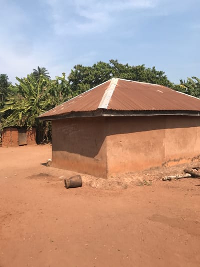 A weathered, single-story building with a corrugated metal roof sits on a dusty, reddish-brown ground. The walls are a muted, earthy tone. Tropical vegetation, including banana trees and tall trees, borders the structure. A small, cylindrical object rests on the ground near the building. The sky is visible above the trees, displaying a pale blue hue with scattered clouds.