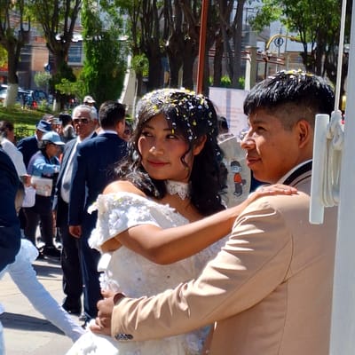 In Cerro Colorado, Peru, a newlywed couple, the bride adorned with confetti in her dark hair and a white lace-trimmed dress, and the groom in a tan suit, are seen posing for photographs amidst a crowd of onlookers. The setting appears to be outdoors, possibly in a public square or park lined with trees. A building is visible in the background. A white rope wrapped around a white plastic tube is visible above the couple.
