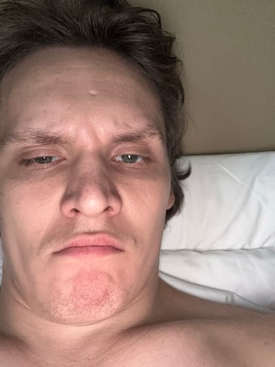A close-up, low-angle selfie shows the face of a person lying down in bed, looking upwards at the camera. The person has light skin, dark brown hair that is slightly messy, and light green eyes. Their expression is neutral to slightly tired. A white pillow is visible behind the person's head and shoulder. The background appears to be a wall with a textured surface. The image was taken indoors, likely in a bedroom. San Francisco, United States of America is not depicted in the image.