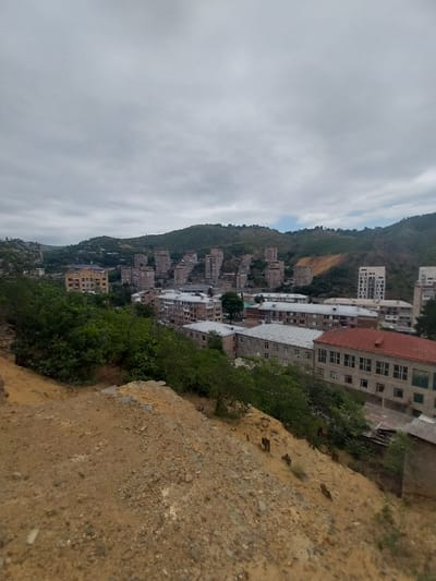 A slightly elevated, wide-angle view captures a dense, urban landscape nestled against a backdrop of green, rolling hills under a cloudy sky. In the foreground, a steep, sandy embankment with sparse vegetation slopes downwards towards the city. The middle ground is dominated by a collection of mid-rise apartment buildings, predominantly constructed of concrete and displaying a variety of architectural styles, some appearing older and more weathered than others. One building with a red tiled roof stands out prominently. The buildings are tightly packed, creating a sense of density within the city. The background reveals further hills, some of which show signs of human intervention, such as cleared areas and construction. The overall impression is of a somewhat overcast day in a hilly, urban environment in Kapan, Armenia, likely a residential district. There are no people or discernible activities visible in the image.