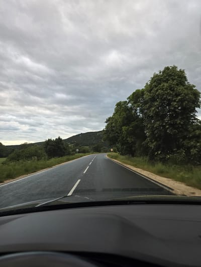 The image depicts a view from inside a vehicle traveling on a two-lane road. The road is paved and features a solid white line on the right shoulder and broken white lines indicating lane division. The landscape to the right of the road consists of green vegetation, including trees and grassy areas. To the left, a cloudy sky is visible. There is a hill or mountain in the distance covered in trees. The image is taken from inside a car, with a portion of the car's interior visible on the right side of the frame. The exact location is Vresovo, Bulgaria.
