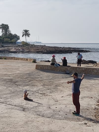 A concrete boardwalk in Rotes, Spain, is seen with a group of people. A man in a blue t-shirt and maroon pants with a cap is standing on the right, arms outstretched. In the foreground a baby or small child sits on the boardwalk. Further away, three women are sitting on the edge of the boardwalk, with one standing in the water. People are visible on the beach. Palm trees are visible and other vegetation near the beach. There are some boats on the horizon.