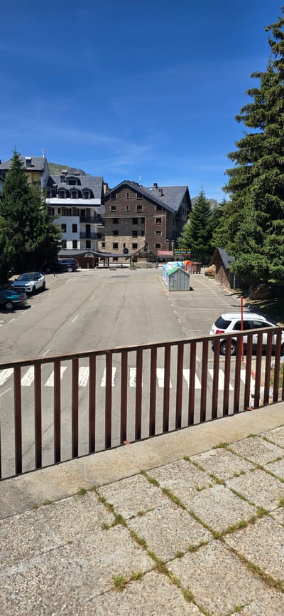 This is a photograph of a street scene in Sallent de Gállego, Spain, likely in a mountainous or resort area. In the foreground, there is a concrete ledge with a wooden railing. Beyond the railing, a paved road with white pedestrian crossing lines is visible. Several cars are parked on the side of the road, including a dark SUV and a white hatchback. On the right side of the road, there are several large, colorful recycling bins and a small wooden structure. In the background, there are multi-story buildings with dark, sloped roofs, typical of alpine architecture. Some of these buildings have stone and wooden facades. Tall pine trees are present throughout the scene, with one particularly large tree on the left and another on the right side of the frame. The sky is a clear, bright blue. No people are visible in the image. The overall impression is of a quiet, sunny day in a mountain town.