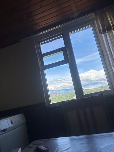 This vertical interior shot depicts a room in Zlatitsa, Bulgaria, looking out through a large window unit. On the left side, the room features a wall or sloped ceiling covered with horizontal wooden panels. At the top, above the window, light-colored, patterned sheer curtains are visible.

The window itself is composed of a large upper pane and a smaller, bottom pane. The smaller pane is partially open, tilting inward. A dark, rectangular fabric item, resembling a temporary blind or sun shade, is attached to the frame of this open window.

Through the window, a bright outdoor scene unfolds under a blue sky dotted with scattered, fluffy white cumulus clouds. Beyond, a verdant, hilly or mountainous landscape is visible, appearing densely vegetated. Some distant buildings or structures, including a hint of a red roof, can be discerned amidst the greenery.

In the bottom right corner of the room, a white, front-loading washing machine is partially visible, showing its control panel with dials and buttons. The brand "AEG" is discernible on its surface. The scene is illuminated by bright daylight, suggesting it is midday or early afternoon. No people or specific activities are depicted.