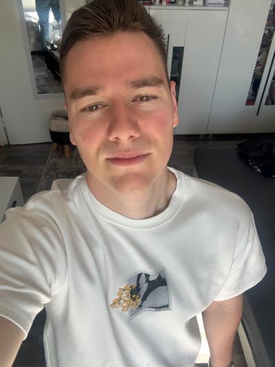 A close-up, indoor selfie depicts a young man with fair skin, brown hair, and brown eyes. He is wearing a white t-shirt with a graphic design on the chest. The background includes a glimpse of a white wardrobe or cabinet, a dark grey sofa, and a partial view of the room's floor, revealing a carpeted surface. The scene appears to be in a residential setting. The location is not discernible as Bukovlak, Bulgaria, based solely on the visual content of the image.