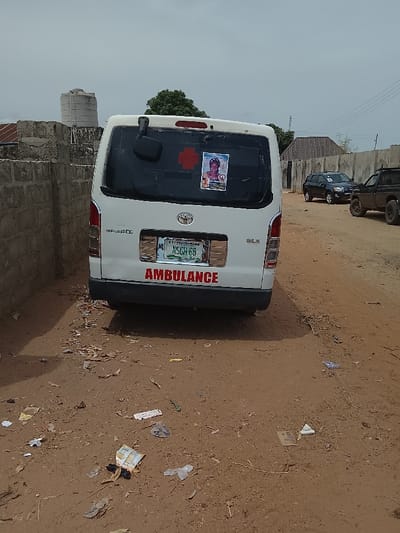 The image shows a white Toyota Hiace ambulance parked on a dirt road in Mukaiya, Nigeria (8.4 latitude, 8.342 longitude). The rear of the vehicle is visible, displaying a red cross emblem and the word "AMBULANCE" in red lettering. The license plate reads "MSCH 68". A small image of a young woman is visible on the rear window. The ground is dusty with some debris.