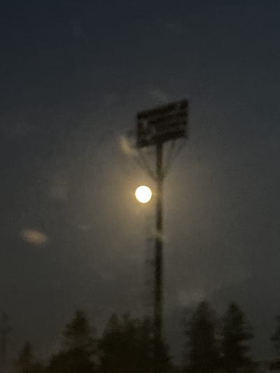 This is a low-light, slightly blurred, full-frame photograph taken in Berezhany, Ukraine. The primary subject is a tall, industrial-style light pole with a rectangular light fixture at its apex. A bright, circular light source, likely the moon or a very bright streetlamp, is positioned behind and slightly to the left of the light fixture. The sky is a dark, hazy gray, with indistinct clouds or atmospheric effects that create soft halos around the light source. In the foreground, silhouetted against the sky and light, are the dark, indistinct shapes of trees and possibly other vegetation. The overall impression is a nocturnal scene with artificial and natural light sources interacting with a dark, natural landscape.