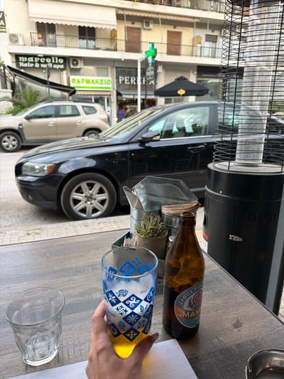 The photograph shows a table at an outdoor cafe in Glifada, Greece, with an overturned glass in a person's hand pouring a yellow liquid into another empty glass. A bottle of Alfa beer is lying on the table. A silver watering can holds small purple flowers. A black car is parked near the table, and another vehicle is visible in the background. Businesses across the street include "Marcelle Hair & Nail Spa" and "ΦΑΡΜΑΚΕΙΟ (Pharmacy) PERFE." A patio heater stands next to the table.