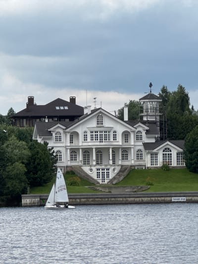 The media file shows a scenic view of a multi-story white building with a dark roof, possibly a house or a hotel, situated on the bank of a body of water. The building features numerous windows, some arched and some rectangular, and has an intricate architectural design with multiple levels and projections. In front of the building, a small sailboat with two sails is moored near the shore, with two people visible onboard. The surrounding area is lush with green trees and grass, suggesting a summer or spring season. The sky is partly cloudy, indicating it might be daytime with potentially variable weather. On the embankment, a rectangular sign with text is visible. The text on the sign reads "ТИХИЙ ХОД", which translates to "SLOW SPEED" in Russian. The scene evokes a sense of tranquility and leisure, hinting at a recreational area or a peaceful residential setting. The overall atmosphere is serene and picturesque, with the water reflecting the sky and surrounding greenery.