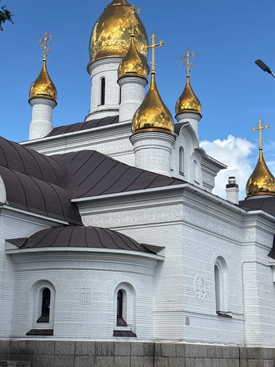 This media file depicts a close-up, angled view of the exterior of a white brick church with golden domes. The church is a prominent subject, with its ornate architecture and shimmering domes dominating the frame. Several golden domes are visible, each topped with a cross. The white brick walls are textured and feature decorative carvings and arched windows. Dark brown, segmented roofs are visible between the domes and the main body of the church. The sky is a clear, bright blue, with a few white clouds present, suggesting fair weather. A street lamp or light fixture is visible in the upper portion of the frame, silhouetted against the sky.
The scene is likely set during the daytime, given the bright lighting and blue sky. The architecture strongly suggests a Russian Orthodox church. While there are no people or overt activities depicted, the image captures the grandeur and religious significance of the building. The focus is on the structural and decorative elements of the church itself, set against a serene sky. No text is visible in the image.