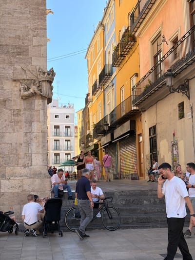 The image depicts a street scene in Alboraia, Spain, with a focus on a section of the historic architecture and a lively pedestrian area. On the left, a weathered stone building, possibly part of a church or ancient structure, dominates the foreground with a sculpted frieze featuring angelic figures and a crown. To the right, a row of brightly colored buildings, predominantly yellow and peach, line a street with balconies. Numerous people are visible, engaged in various activities. Some are seated on stone steps, others are walking or standing. A man in a white t-shirt and black pants is walking towards the right side of the frame, holding a phone to his ear. A bicycle is parked near the steps. In the background, more buildings and people are discernible, suggesting a bustling urban environment. The sky is clear blue.
