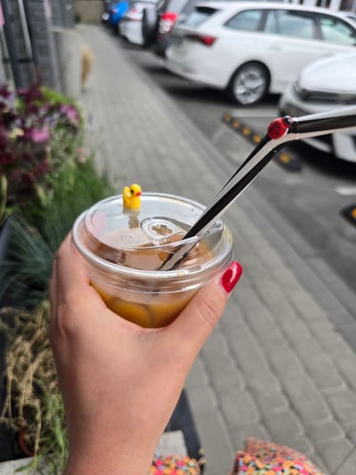 A person's hand with red-painted fingernails holds a clear plastic cup with a yellow drink and a lid. A black and white striped straw is inserted into the lid, decorated with a red ladybug charm and a small yellow duck figure. The cup is positioned over a blurry background of green plants and a grey brick wall. In the upper left, a blurry white car is visible. The image appears to have been taken outdoors during daylight. The lighting suggests it might be a sunny day. The setting seems to be urban or suburban, possibly a cafe or a street scene. The overall impression is casual, with a focus on the beverage and its playful decorations. The hand and cup are in sharp focus, while the background elements are intentionally blurred, drawing attention to the drink.