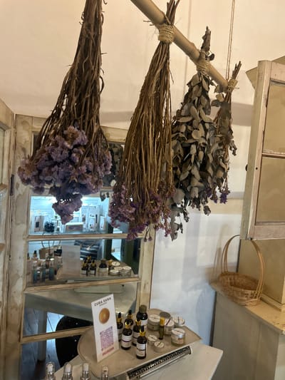 The image shows a shop interior in Escazú, Costa Rica, likely a natural beauty products store. Bundles of dried flowers and leaves hang from a wooden rod on the ceiling. These appear to be for sale or decorative. In the background, a mirrored, wooden cabinet displays a range of small bottles and jars of various sizes, presumably the store's products. In the foreground, a wooden counter displays more product jars and bottles, alongside a QR code sticker with the words “CUBA SANA” and “HACIENDA,” likely brand information. The overall environment is clean and well-lit. The store's interior is decorated with a rustic theme, featuring white-painted walls, distressed wooden furniture, and wicker baskets. The image captures a moment of calm and tranquility in a small business setting.