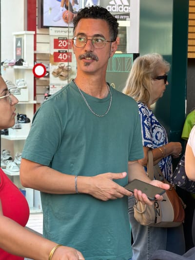The image captures a medium shot of a man with dark, curly hair and a mustache standing in front of a store in Ashdod, Israel. He is wearing a teal t-shirt, glasses, and a silver chain necklace. The man is holding a black smartphone in his right hand. To his left, a woman in a red top is partially visible. In the background, shelves display shoes, and promotional signs with numbers are visible, suggesting a retail environment. A woman with blonde hair wearing sunglasses and a blue and white patterned shirt is also in the background, as is a beige and brown patterned bag. The overall scene depicts a casual moment in a commercial setting.