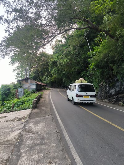 In the city of Meudheun, Indonesia, a white van is captured driving on a winding asphalt road bordered by lush green foliage and rocky inclines. The van, loaded with several sacks on its roof, displays a license plate that reads "BG 1121 EZ." To the left of the road, a small, weathered wooden structure with a corrugated metal roof stands amidst dense vegetation. The foreground features a concrete walkway that transitions into a rougher, more textured path. The scene is bathed in natural light, suggesting it is daytime.