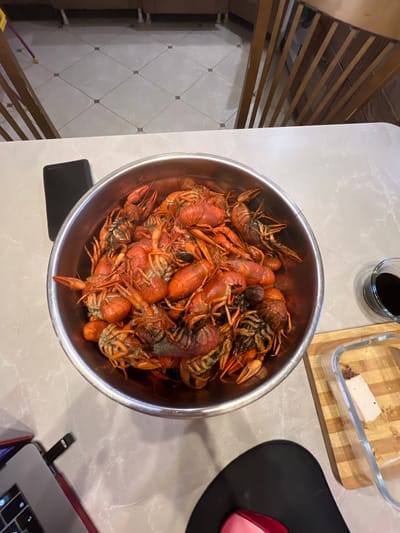 A close-up, overhead shot captures a large metal bowl overflowing with cooked crawfish, placed on a light-colored, speckled countertop. The crawfish are a vibrant red-orange, with many tails curled and claws interlocked. To the right of the bowl, a small glass dish contains a dark liquid, likely a dipping sauce, and a clear glass container with a wooden base holds a piece of what appears to be food, possibly cheese. In the background, a portion of a kitchen island with wooden chairs is visible, with a tiled floor in the distance. On the left side of the image, a black smartphone rests near the bowl, and further left, a laptop computer with a red case is partially visible. The scene is situated on a countertop, suggesting an indoor setting, potentially a kitchen or dining area. There are no people present in the image. The image does not provide any specific information to place the scene in Bol'shaya Shishovka, Russia.