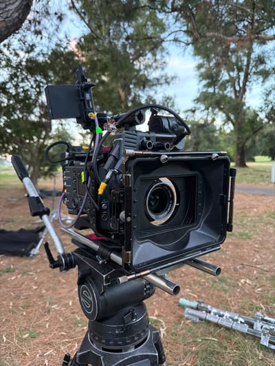 A professional film camera on a tripod is set outdoors. The lens is visible within a matte box. Cables and accessories are attached. The tripod head is black with white lettering. A partially visible, disassembled tripod lies on the ground nearby. Trees are visible in the blurred background, suggesting a park or wooded location. The camera is positioned low to the ground.