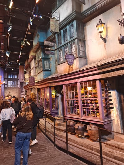 The image depicts a crowded scene in the "Diagon Alley" set at the Warner Bros. Studio Tour London in Watford, United Kingdom. The street is lined with whimsical, leaning buildings, typical of the set design. The storefronts, such as "Mr. Mulpepper's Apothecary" and "Slug & Jiggers Apothecary," are visible, with their windows displaying various curious items. A line of people is walking along the street, partially obscured by the foreground. A metal railing separates the walkway from the raised platforms in front of the storefronts. Above, the ceiling is exposed, showing lighting rigs and other equipment. The lighting creates a warm, inviting atmosphere, enhancing the immersive experience of the set.