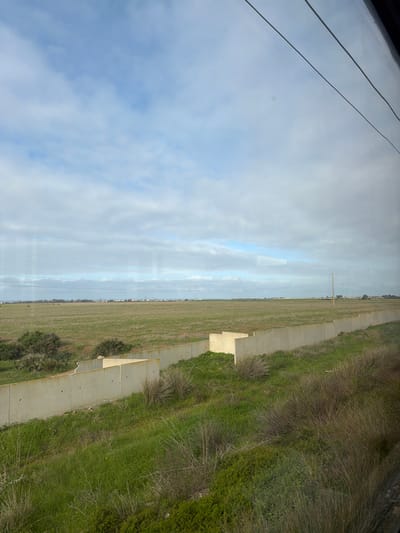 This image presents a wide view of a rural landscape within El Mansouria, Morocco, observed from an elevated vantage point, likely through a reflective window. The upper frame consists of a partially overcast sky with light blue areas interspersed with white and grey clouds. Two dark, thin overhead wires are visible in the top right. A light grey concrete barrier, constructed from interlocking panels and featuring corner sections, traverses the midground horizontally. Below this barrier, the immediate foreground is a green grassy slope with patches of dry, bushy vegetation. Beyond the barrier, a large, flat expanse of green field extends to the distant horizon. Along the horizon line, a faint array of structures or buildings indicates a remote settlement or industrial zone. A single wooden utility pole with cross-arms stands in the field towards the right side of the frame. No individuals, overt actions, or interactions are present.
